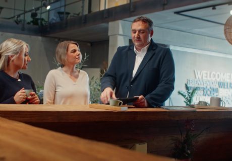 Zwei Frauen und ein Mann stehen mit Kaffee an einer Theke im Eingangsbereich eines großen Gebäudes und reden miteinander.