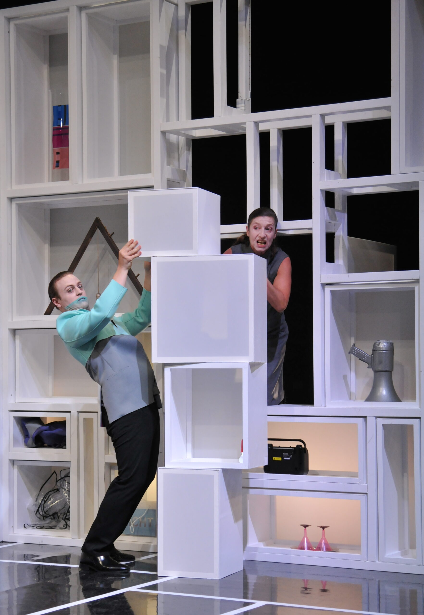 Bühnenbild und Kostümbild Theater – Theaterstück „Doctor Faustus Lights The Lights“, schräg gekleideter Mann stapelt weiße Boxen aufeinander, dahinter blickt eine Frau hervor