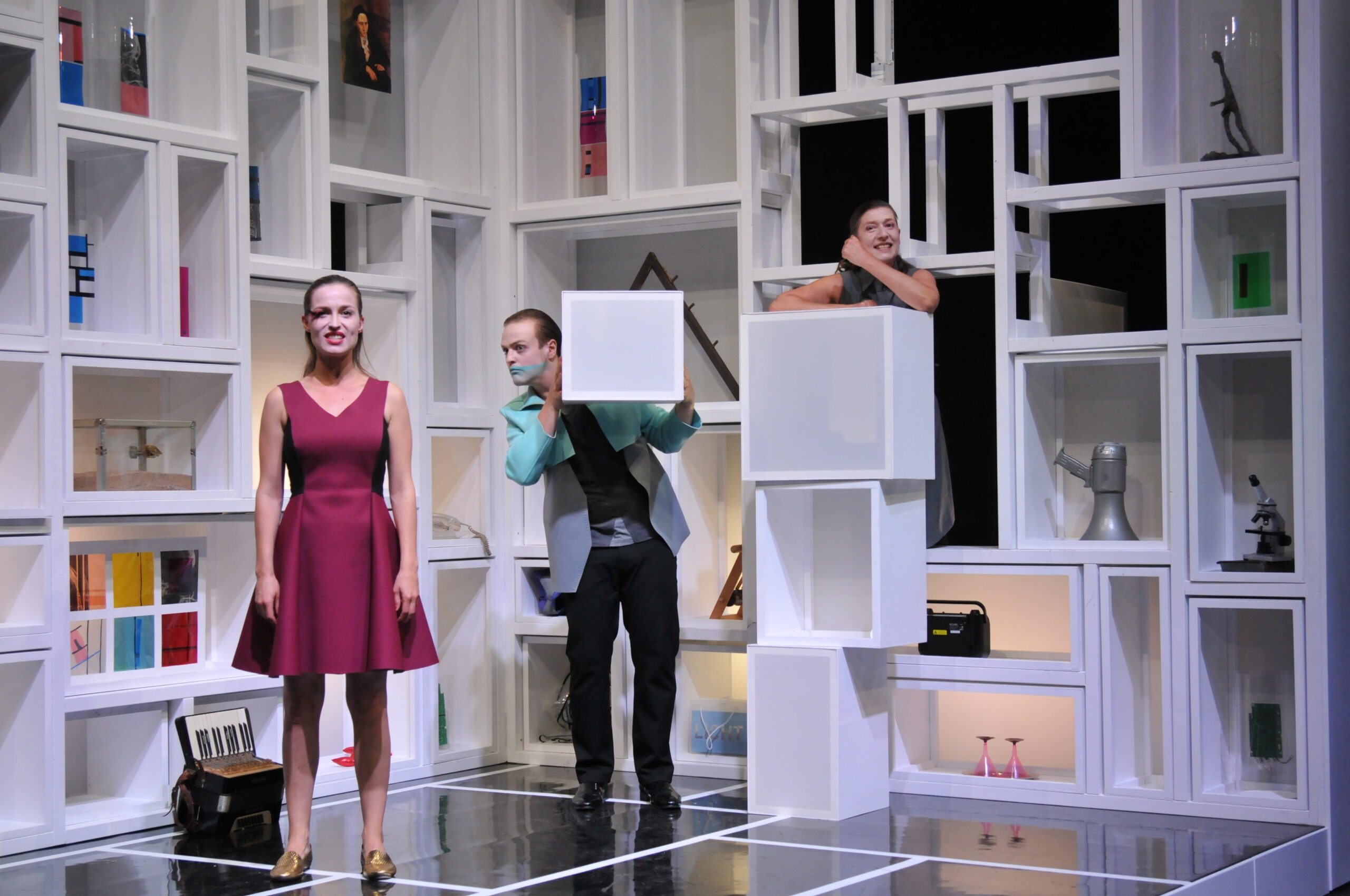Bühnenbild und Kostümbild Theater – Theaterstück „Doctor Faustus Lights The Lights“, schräge Szene – eine Frau in rotem Kleid im Vordergrund, ein Mann blickt neben einer weißen Box, die er selbst in der Hand hält, vorbei, eine Frau schaut über drei gestapelte Boxen.