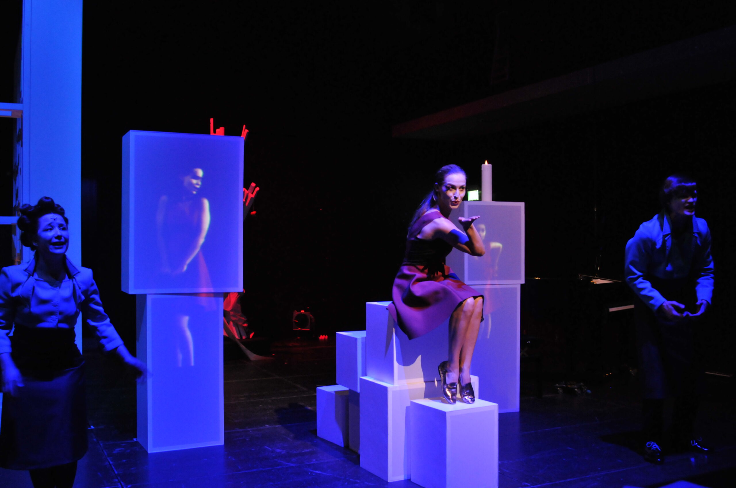 Bühnenbild und Kostümbild Theater – Theaterstück „Doctor Faustus Lights The Lights“, junge Frau sitzt auf einer Konstruktion aus weißen Boxen und macht die Kusshand, Neben ihr zwei Installationen mit Prjektionen auf denen sie auch zu sehen ist.