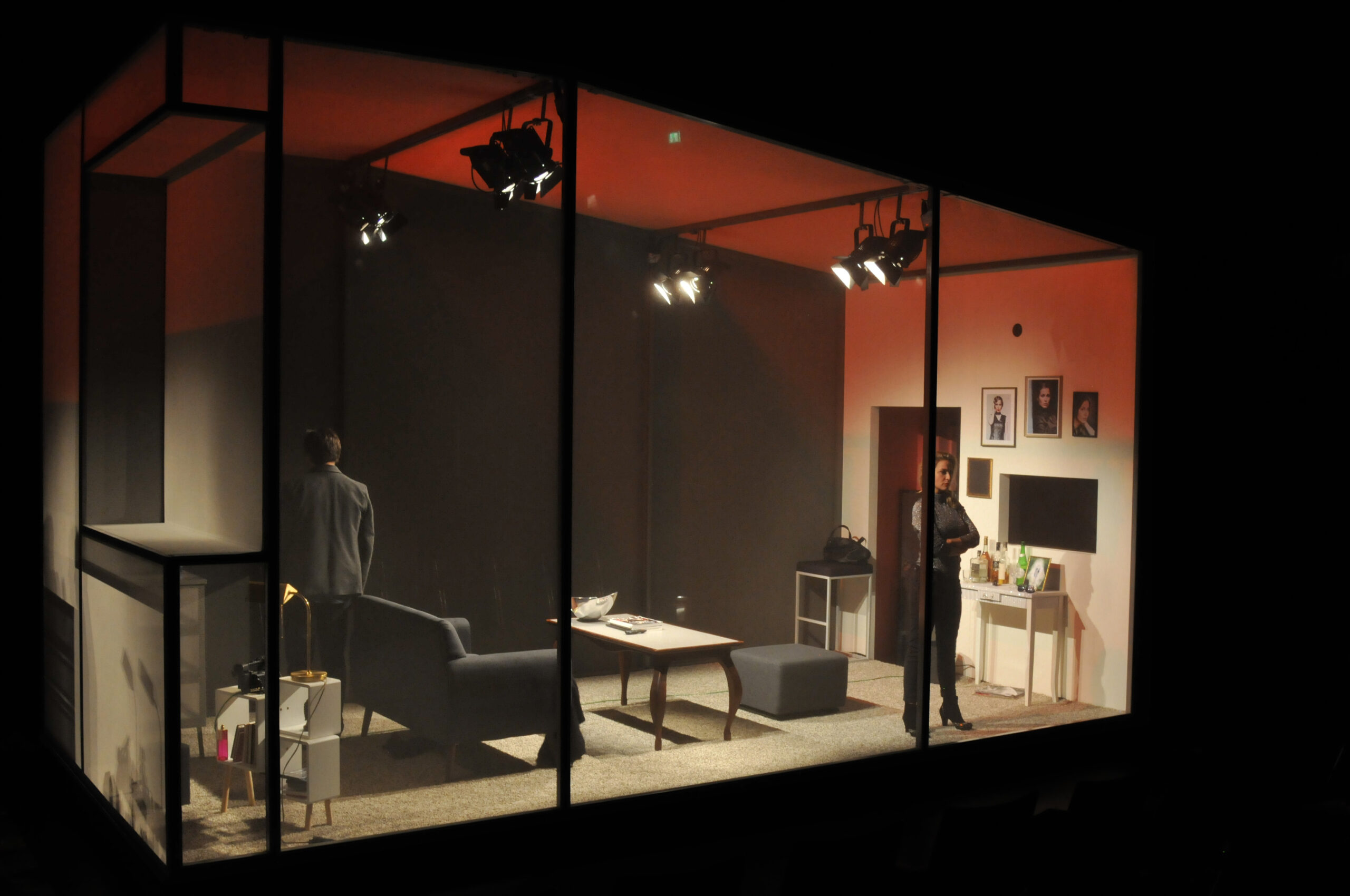 Bühnenbild und Kostümbild Theater – Theaterstück „Das Interview", Blick auf einen durch transparente Wände abgeschlossenen Bühnenraum, in dem sich ein Wohnzimmer befindet. Eine Frau steht an einer Wand
