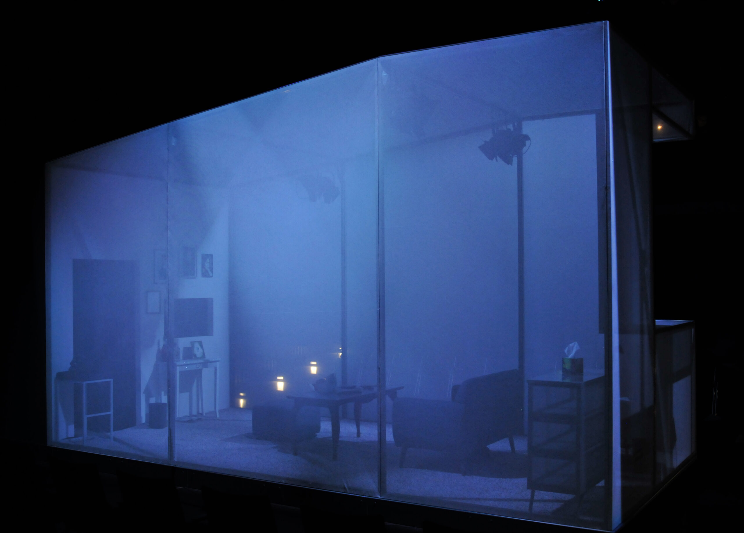 Blick auf einen durch transparente Wände abgeschlossenen Bühnenraum, in dem sich ein Wohnzimmer befindet. Der Raum ist blau beleuchtet.