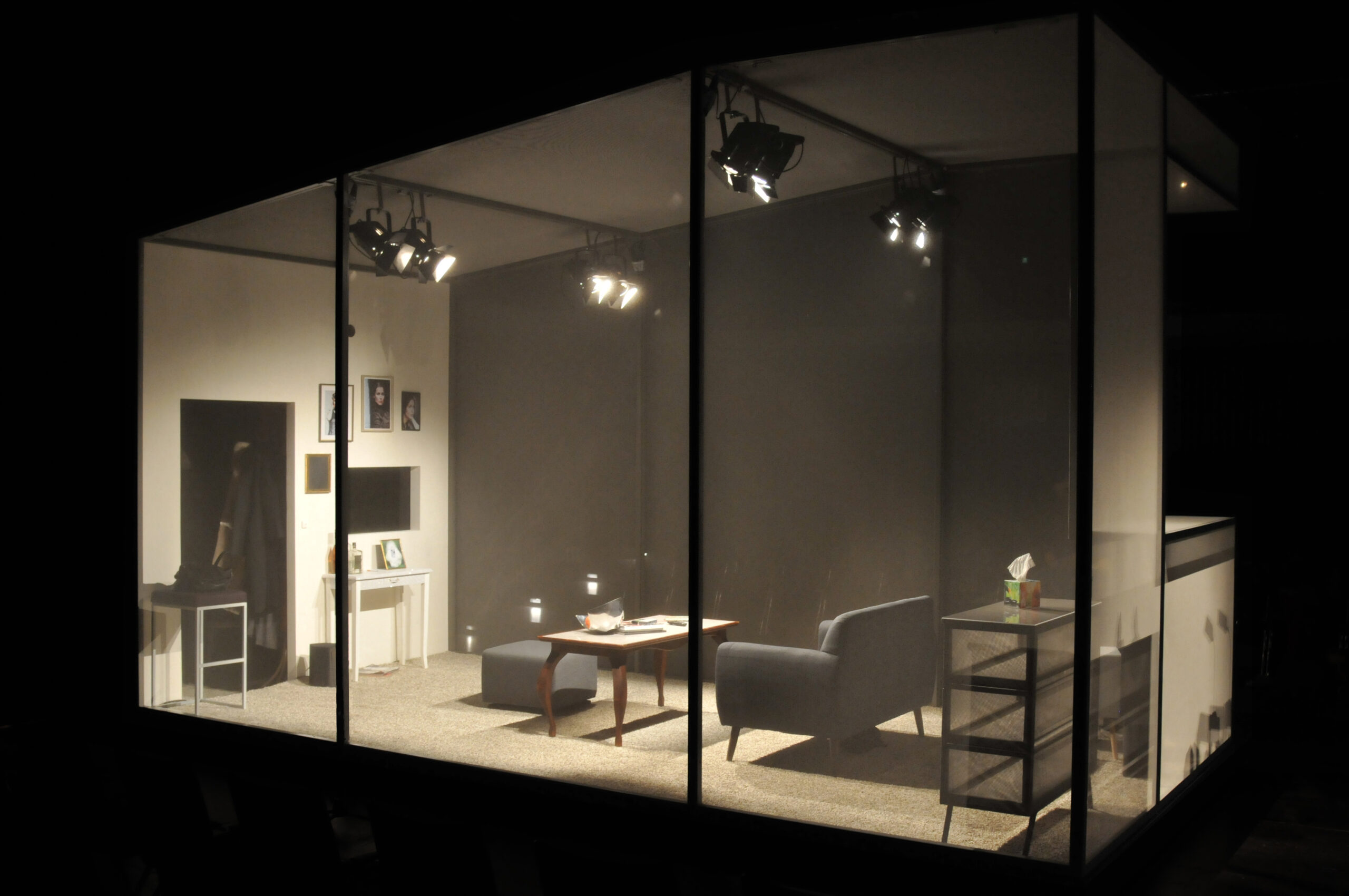 Blick auf einen durch transparente Wände abgeschlossenen Bühnenraum, in dem sich ein Wohnzimmer befindet. Der Raum ist blau beleuchtet.