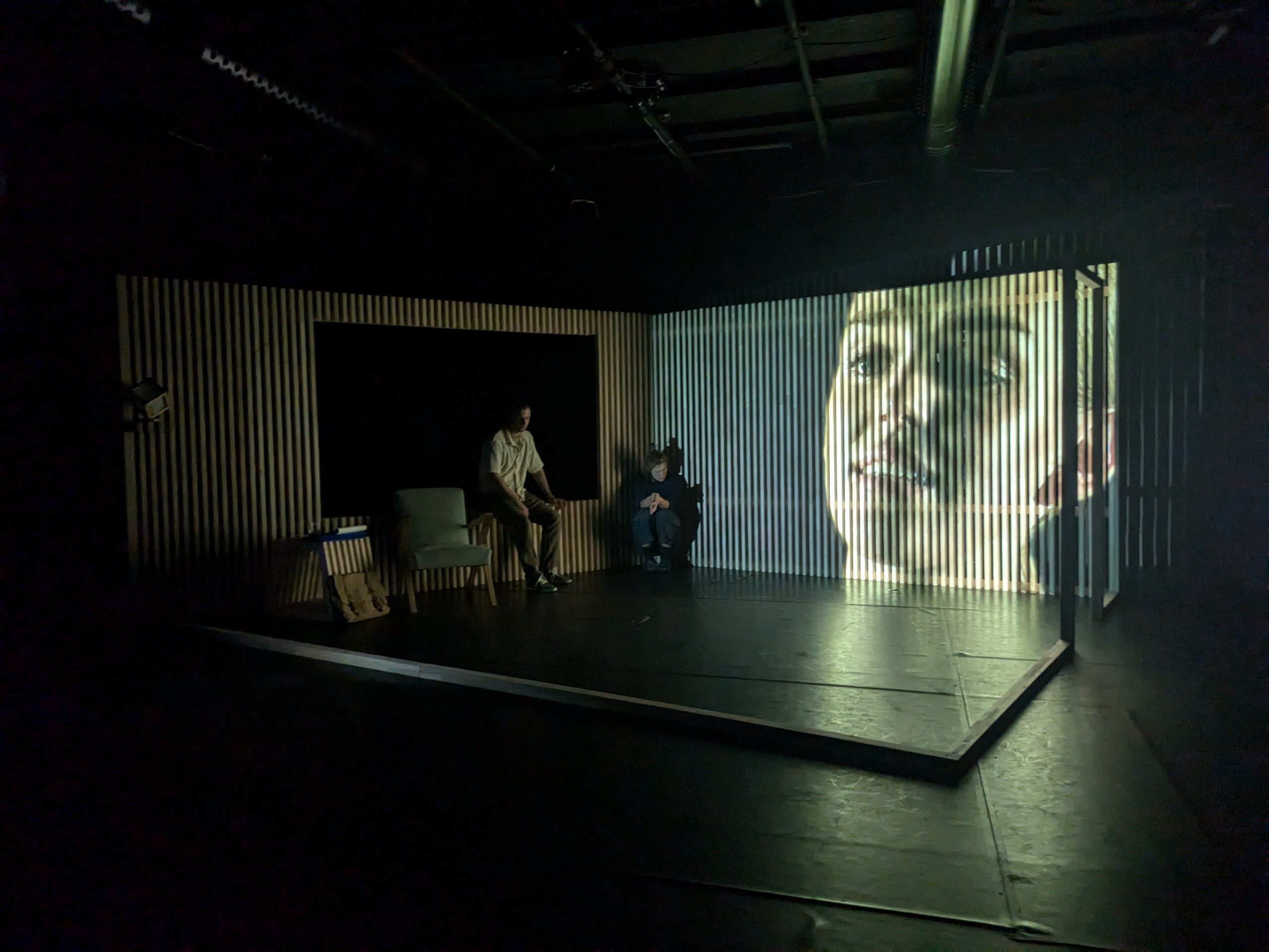 Ein reduzierter Bühnenraum, in dem sich zwei Personen aufhalten, mit einer Hinterwand aus Latten, auf die das Gesicht einer jungen Frau projiziert ist