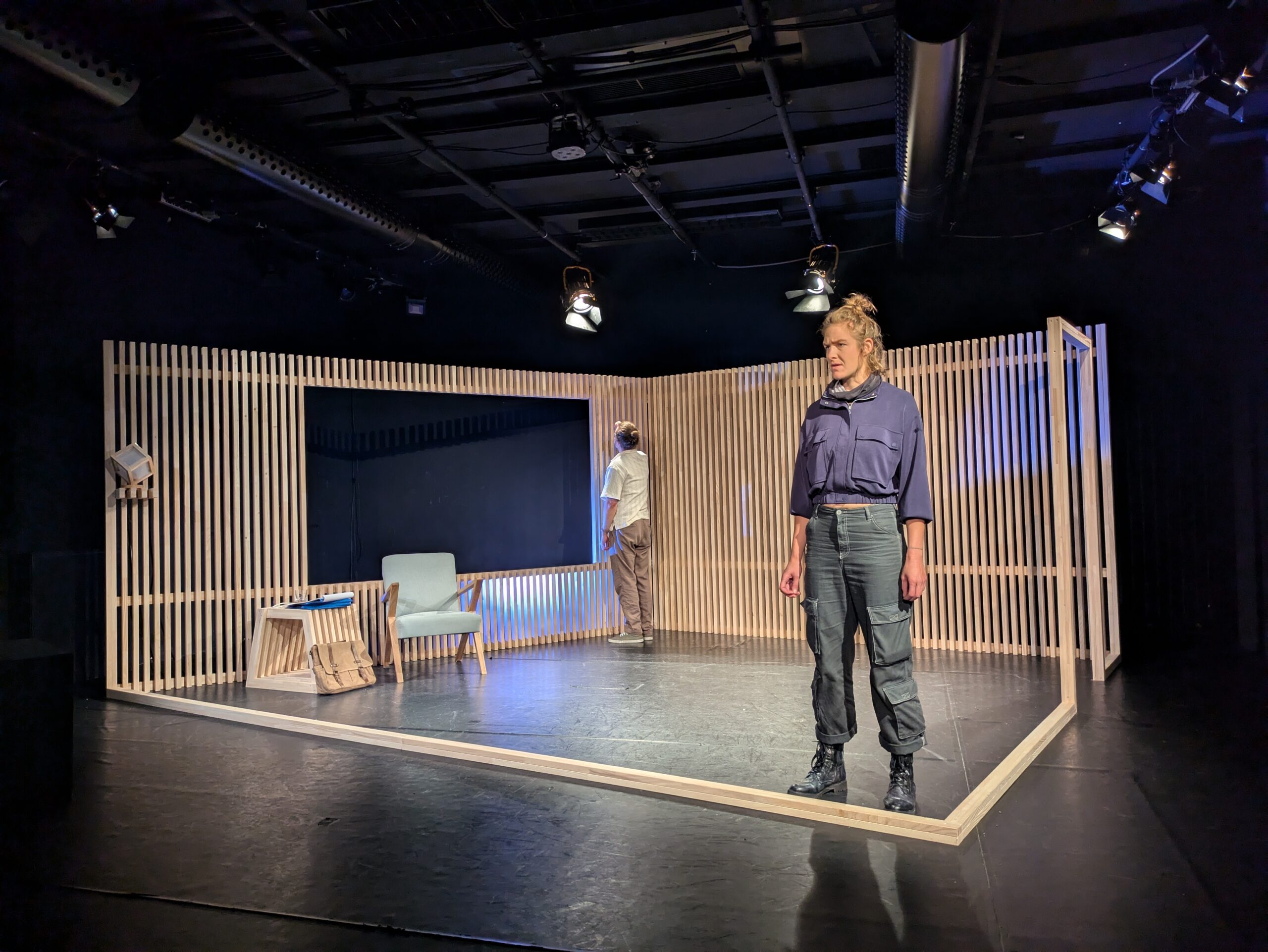 Bühnenbild und Kostümbild Theater – Theaterstück „Freie Wahl“, ein reduzierter Bühnenraum, der ein Zimmer andeutet, im Vordergrund eine junge Frau in dunkler Kleidung, in der hinteren Ecke ein Mann