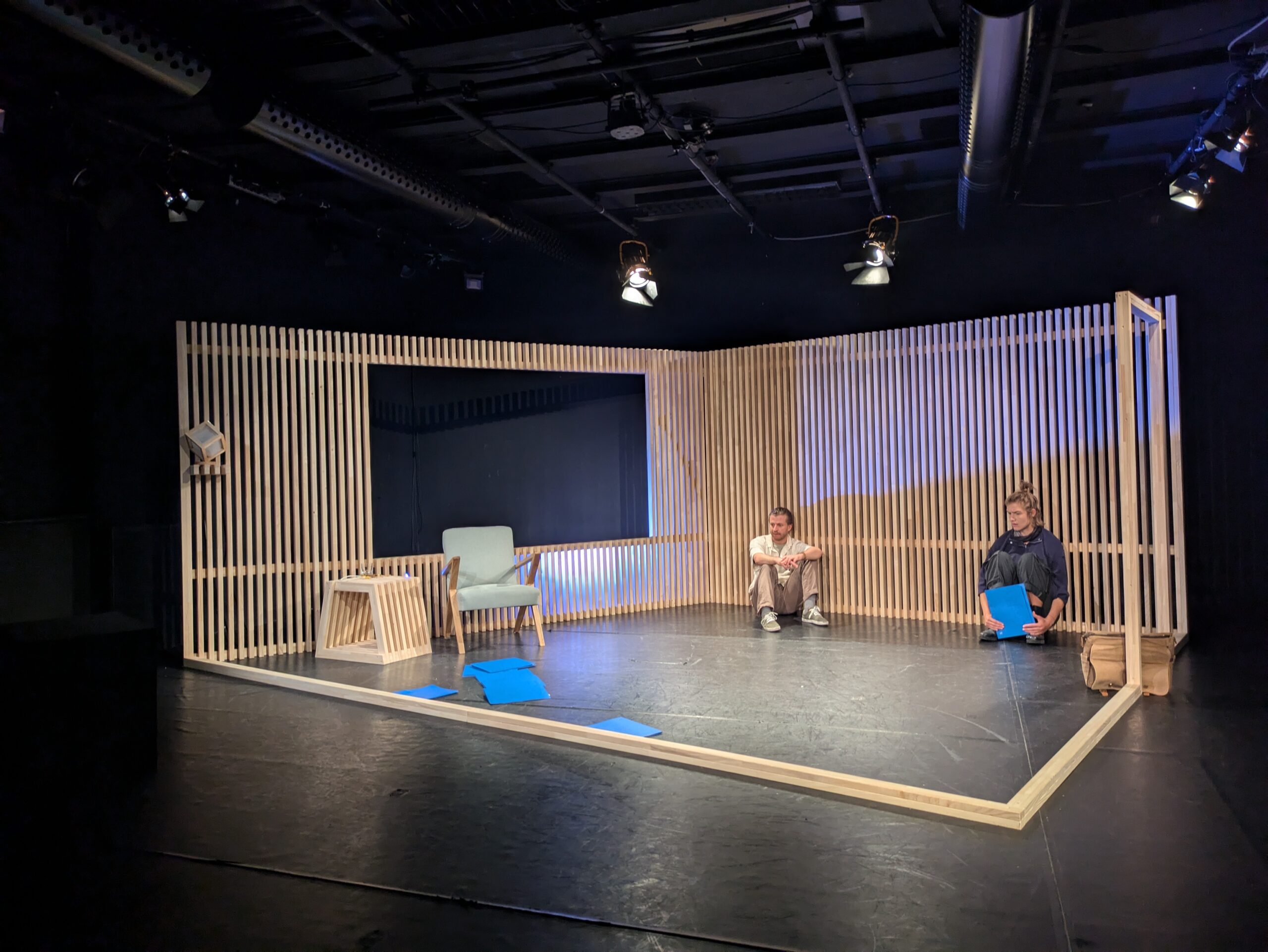Ein reduzierter Bühnenraum, der ein Zimmer andeutet – an der Hinterwand hocken zwei Personen