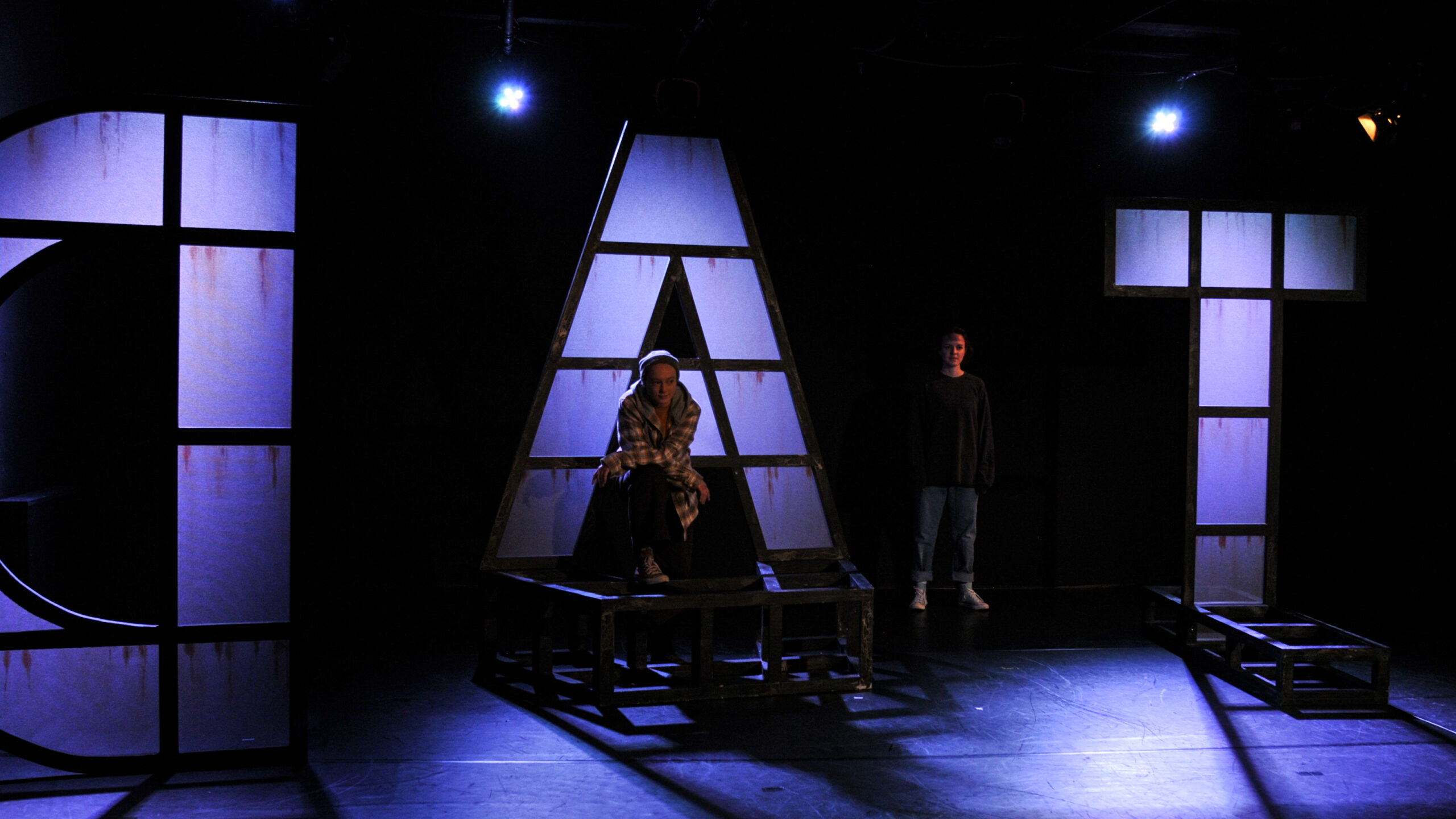 Bühnenbild und Kostümbild Theater – Theaterstück „Das Gesetz der Schwerkraft“, leuchtende große Buchstaben auf einer dunklen Bühne und die Schemen von zwei Personen