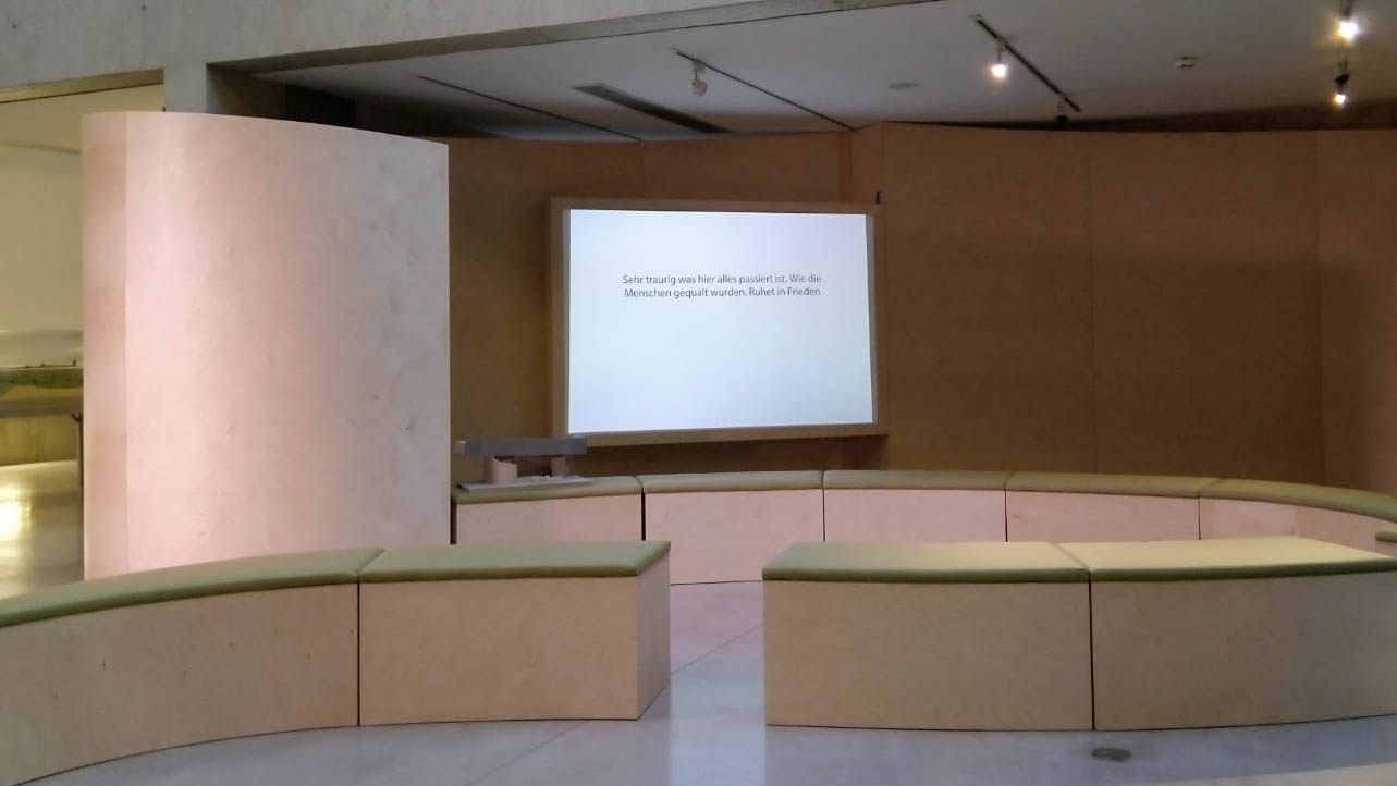 Ausstellungsgestaltung Raumdesign – Design Rauminstallation „mit:teilen“, Ansicht der Rauminstallation: organische Form mit Sitzgelegenheiten mit grüner Polsterung, die vor einem Bildschirm positioniert sind.