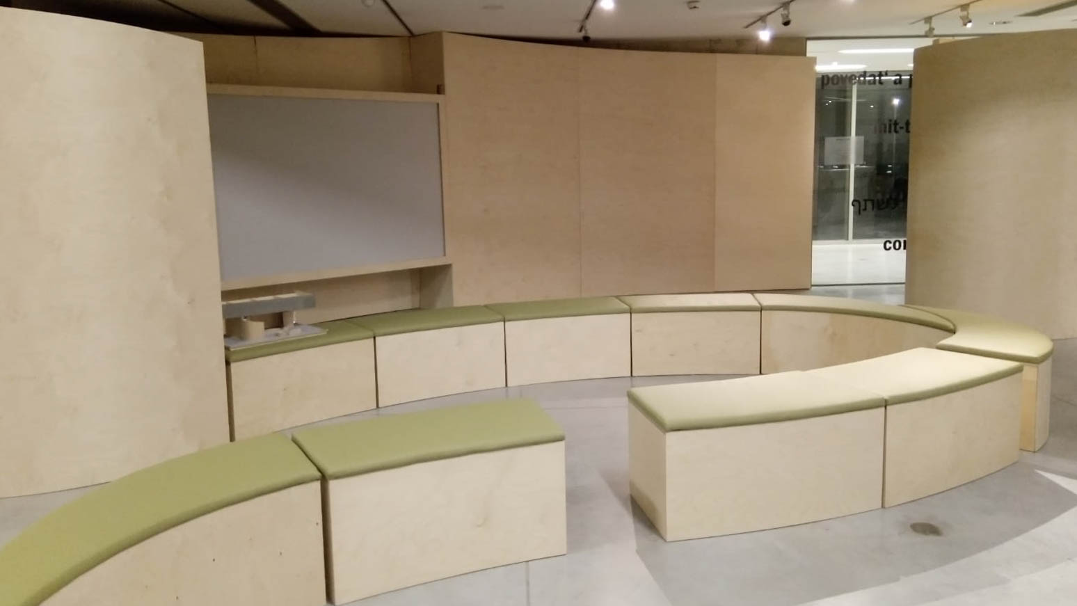 Ausstellungsgestaltung Raumdesign – Design Rauminstallation „mit:teilen“, Ansicht der Rauminstallation: organische Form mit Sitzgelegenheiten mit grüner Polsterung, die vor einem Bildschirm positioniert sind.