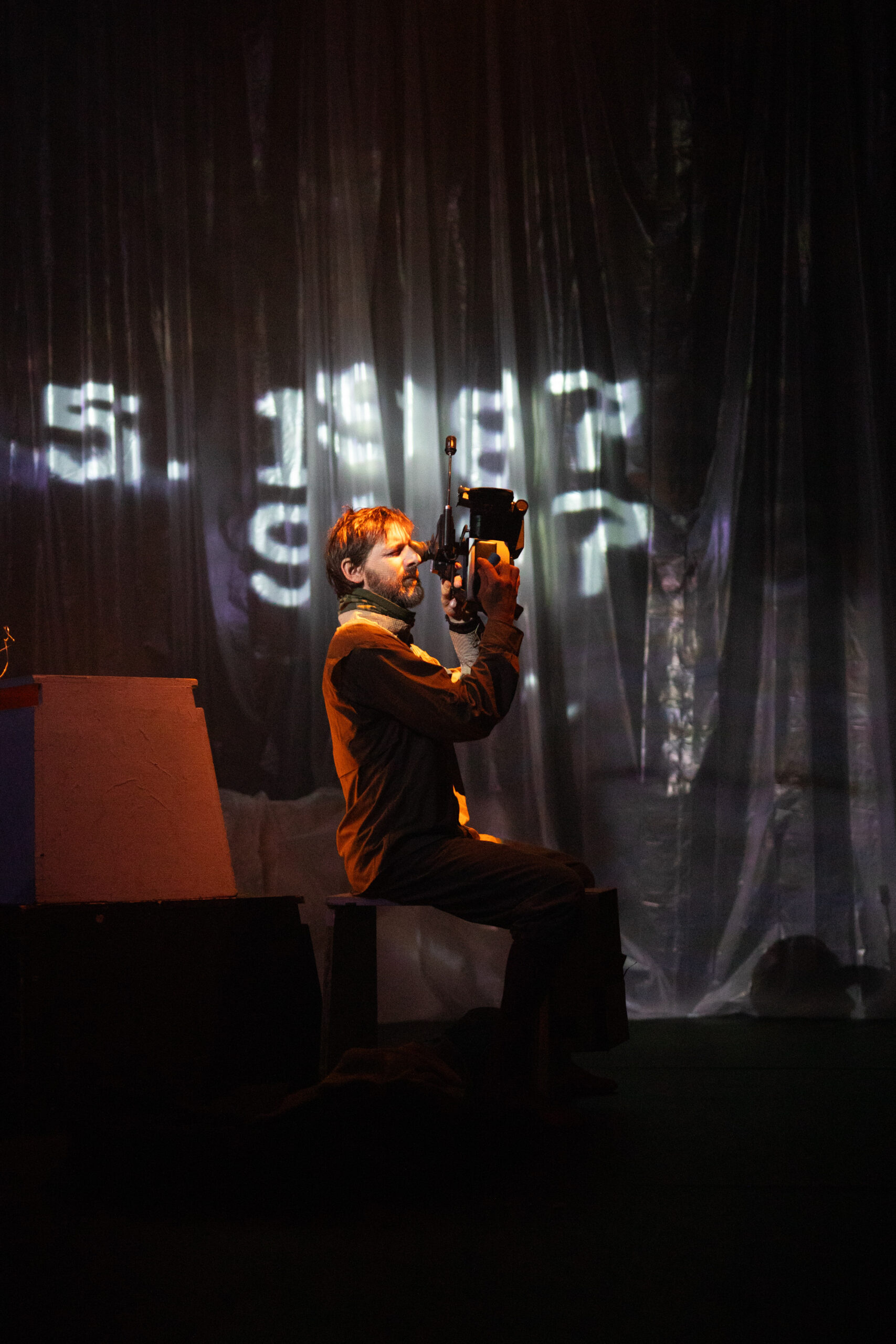 Bühnenbild und Kostümbild Theater – Tanztheater „The Collapse“, ein Mann sitzt mit einer Kamera in der Hand auf einer Box, im Hintergrund Projektionen von Zahlen auf einem Vorhang