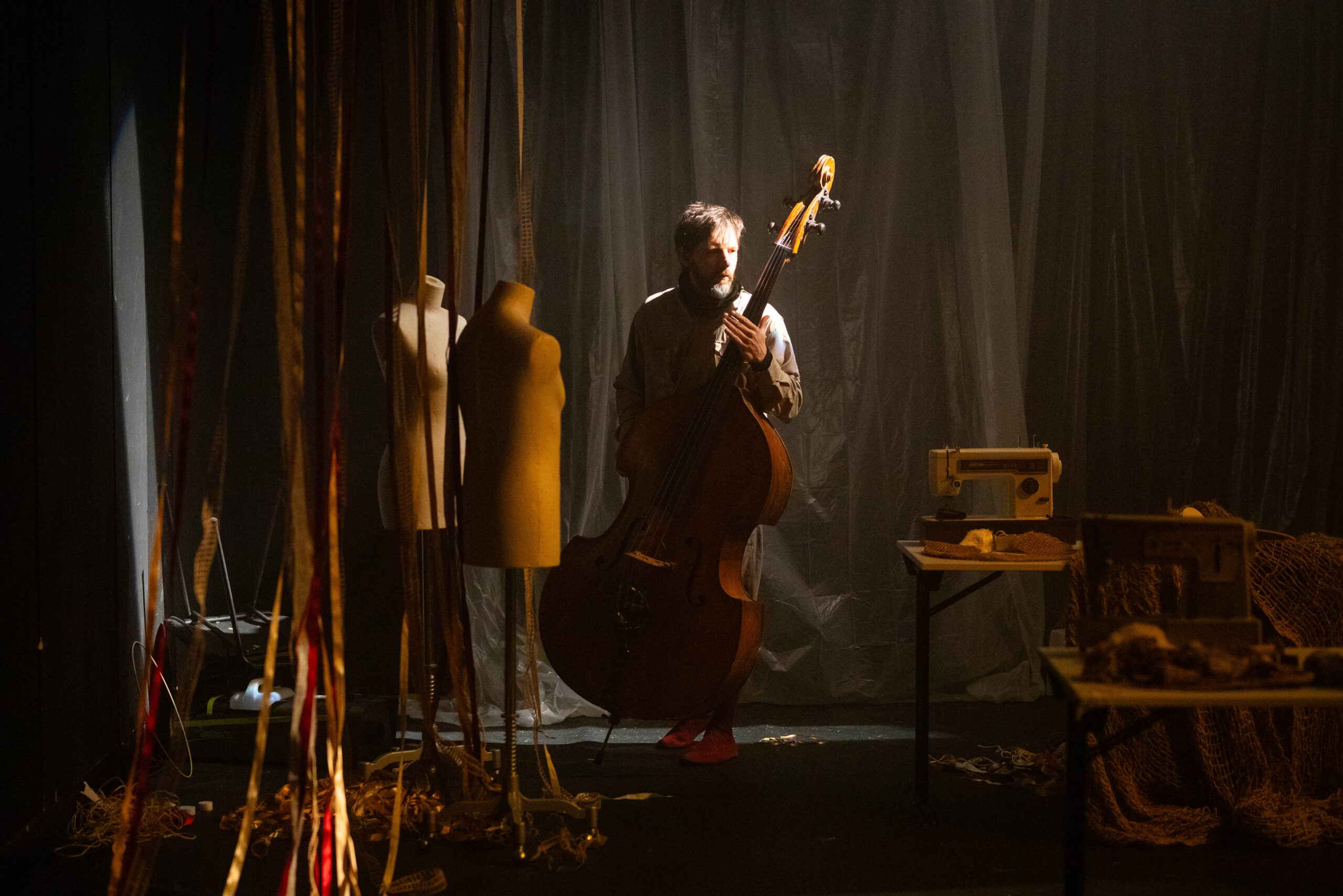 Bühnenbild und Kostümbild Theater – Tanztheater „The Collapse“, ein Mann mit Kontrabass in der Hand geht durch eine dystopische Schneiderwerkstätte