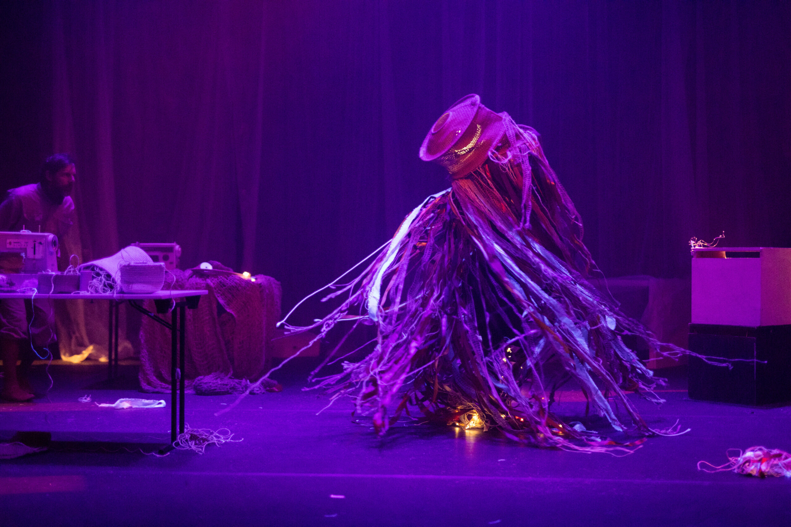 Ein seltsames Wesen, das an Fastnachtsfiguren erinnert, tanzt inmitten einer dystopischen Schneiderwerkstatt