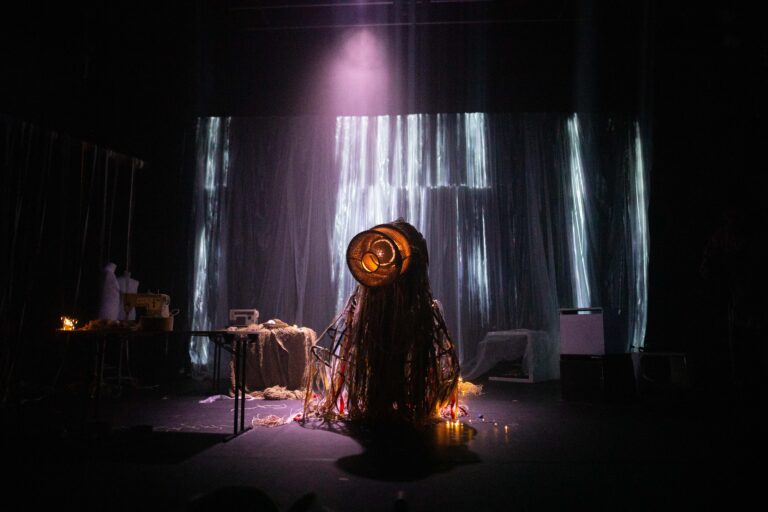 Bühnenbild und Kostümbild Theater – Tanztheater „The Collapse“, ein seltsames Wesen mit langen Fäden am ganzen Körper steht in der Mitte eines dunklen, dystopischen Bühnenraumes