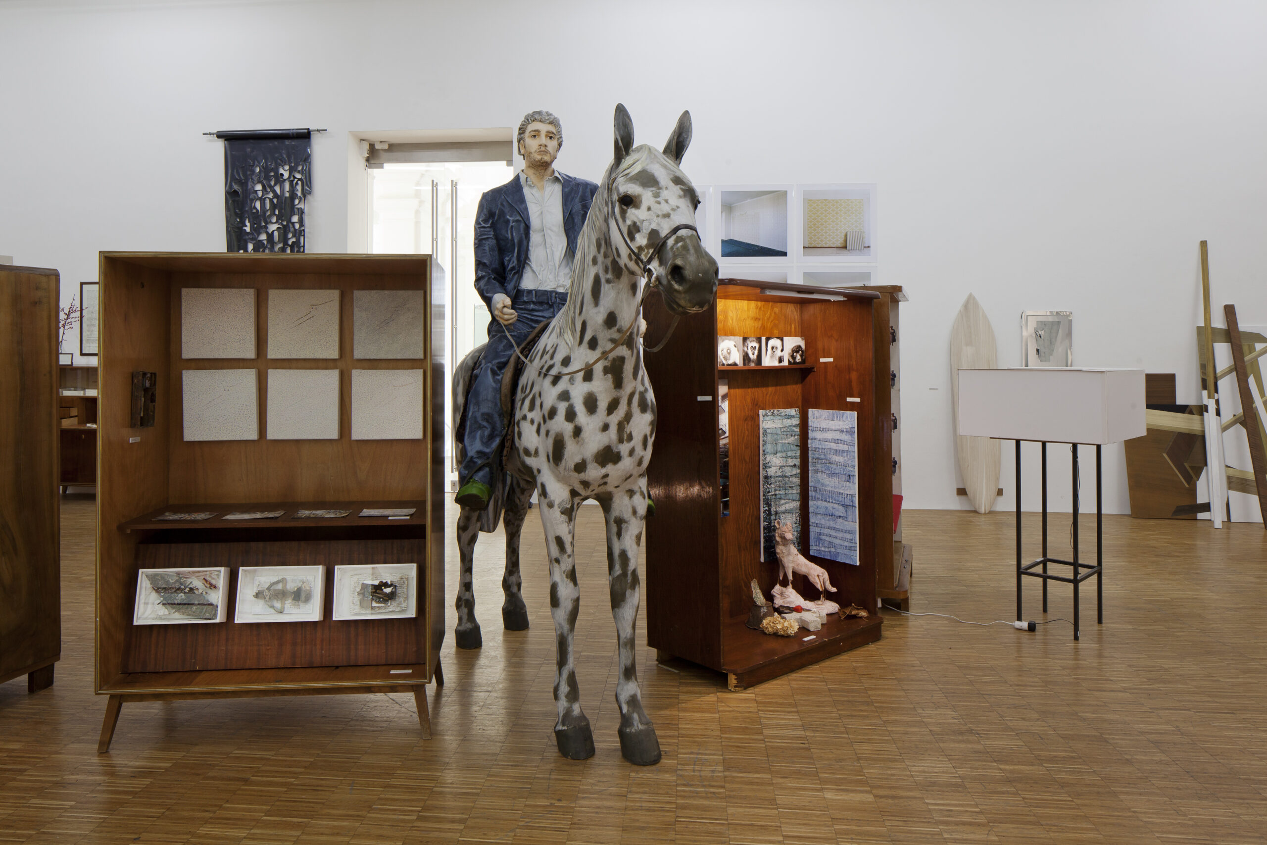 Ausstellungsgestaltung Raumdesign – Design Jahresausstellung „Für die Fülle“, ein Ausstellungsraum wie eine Wunderkammer: ein riesiges Pferd mit Reiter und alte Schränke, in denen sich verschiedene Kunstwerke befinden