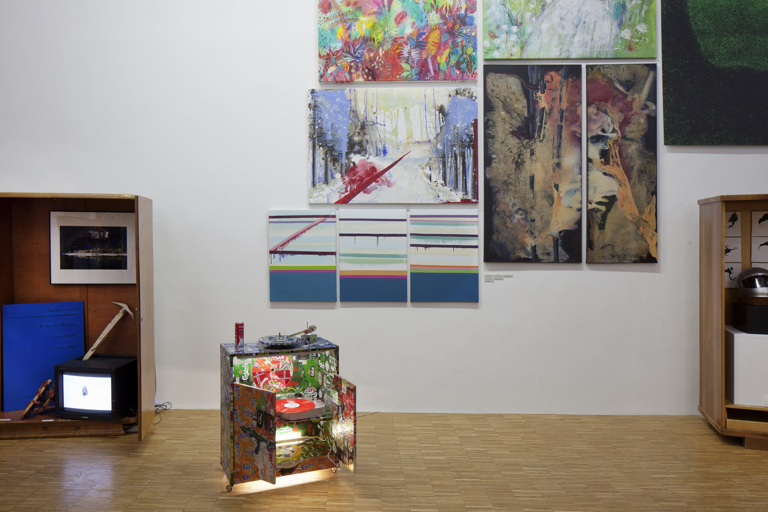 Ausstellungsgestaltung Raumdesign – Design Jahresausstellung „Für die Fülle“, ein Ausstellungsraum wie eine Wunderkammer: eine Wand mit Bildern nebeneinander angeordnet, davor ein beleuchtetes, buntes Schränkchen